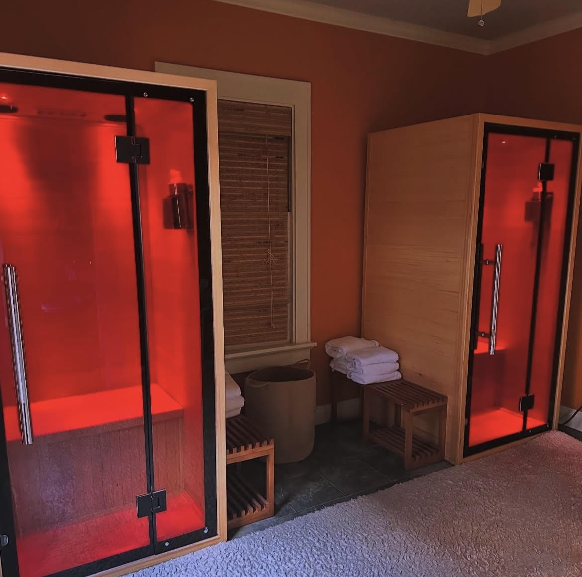 Two infrared sauna rooms with red interior lighting in a room with a window and towels on a stool.